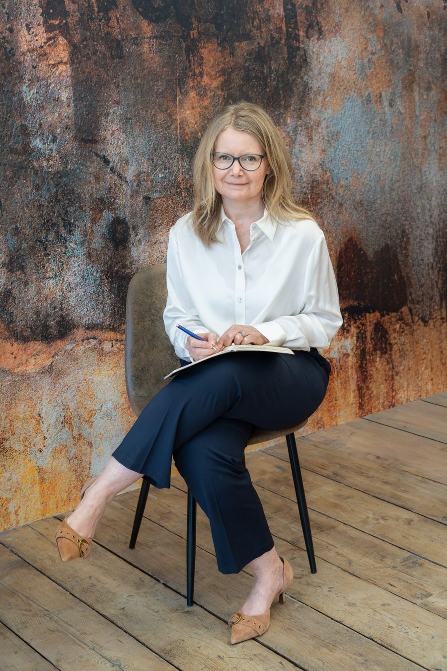 Nicola Pope seated with a notebook in front of a rust-toned wall